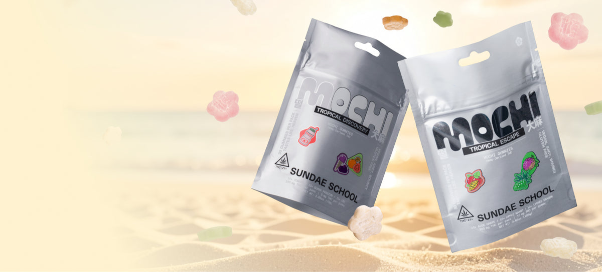 A horizontal image featuring the same two silver "Sundae School Mochi" gummy pouches on a sandy beach setting. The "Tropical Discovery" and "Tropical Escape" pouches are positioned on the right side of the image. Several flower-shaped and rectangular gummies appear to be floating or falling around the pouches. The background shows a soft, out-of-focus beach scene with light-colored sand, calm ocean water, and a bright, hazy sky.