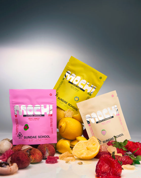 Assorted Sundae School Mochi gummy packs in vibrant colors arranged with fresh fruits like strawberries, grapes, pears, and apples, displayed against a clean gradient background