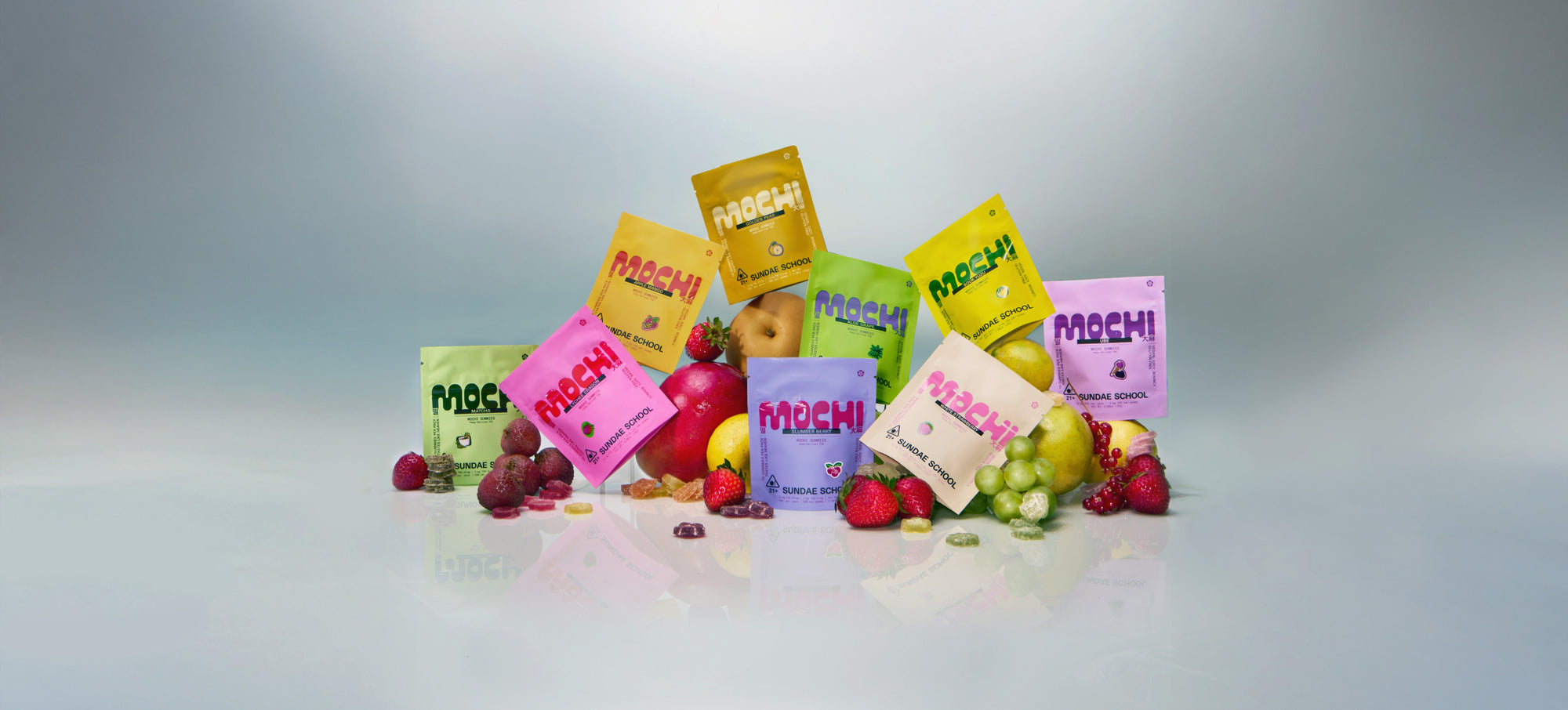 Assorted Sundae School Mochi gummy packs in vibrant colors arranged with fresh fruits like strawberries, grapes, pears, and apples, displayed against a clean gradient background