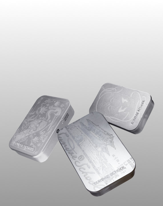 A horizontal web banner showcasing three silver Sundae School tins floating on the right side of the frame, leaving open space on the left. The metallic tins are engraved with artistic designs including a crane, traditional Korean palace architecture, and a tiger, set against a clean gray background.