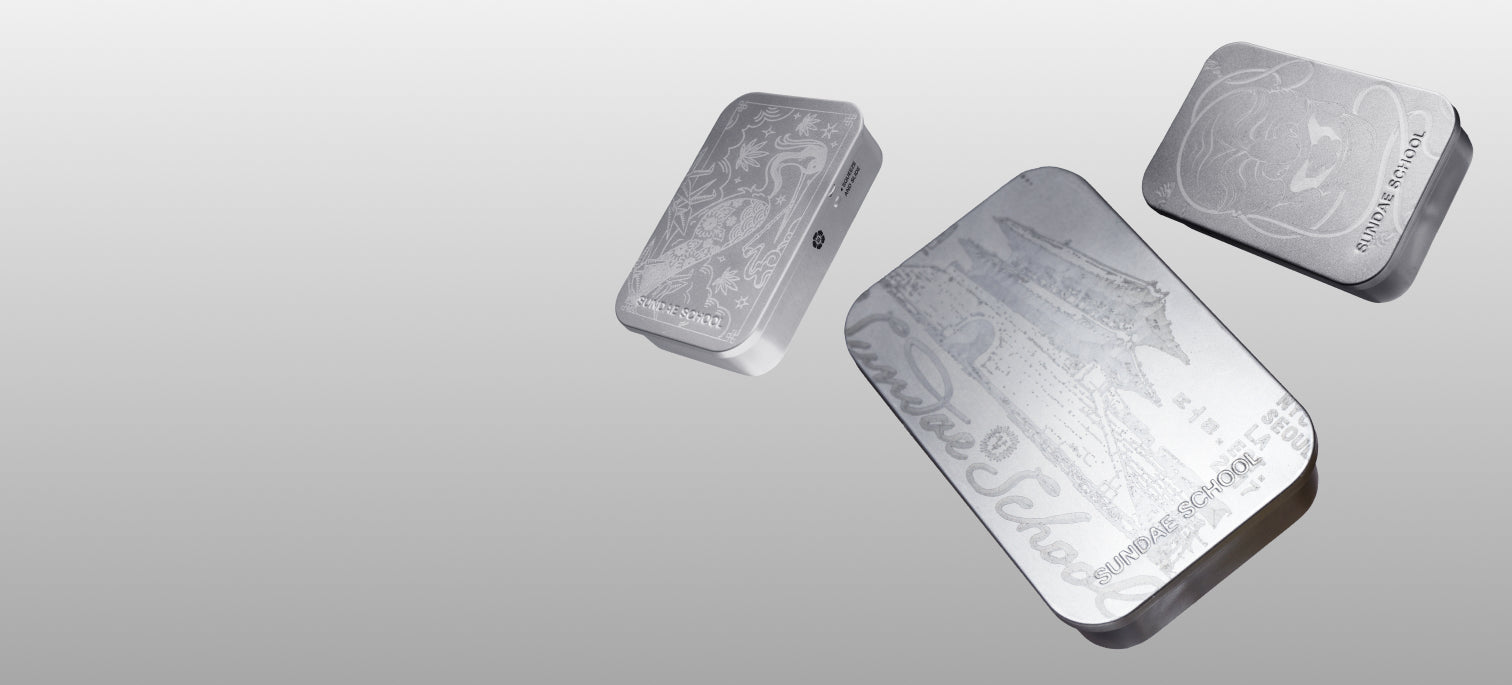 A horizontal web banner showcasing three silver Sundae School tins floating on the right side of the frame, leaving open space on the left. The metallic tins are engraved with artistic designs including a crane, traditional Korean palace architecture, and a tiger, set against a clean gray background.