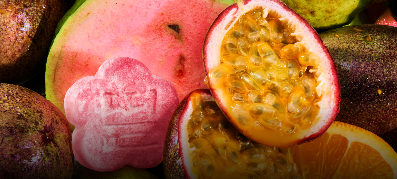 A wide horizontal web banner featuring a macro, close-up shot of fresh tropical fruits, including juicy halved passion fruits with golden pulp and a slice of bright pink guava. A single pink, flower-shaped mochi gummy with a traditional embossed stamp is nestled among the fruit on the left.