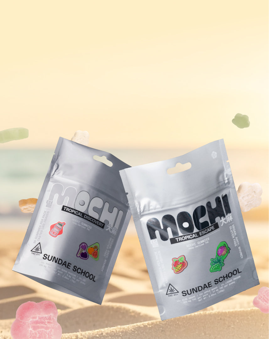A vertical image showing two silver pouches of "Sundae School Mochi" gummies resting on a sandy beach. The pouch on the left is labeled "Tropical Discovery" and features small illustrations of a drink bottle, grapes, and an orange. The pouch on the right is labeled "Tropical Escape" and features illustrations of a strawberry and a dragonfruit. Small, flower-shaped, and rectangular gummies are scattered around the pouches on the sand, with a blurred ocean and sky in the background.