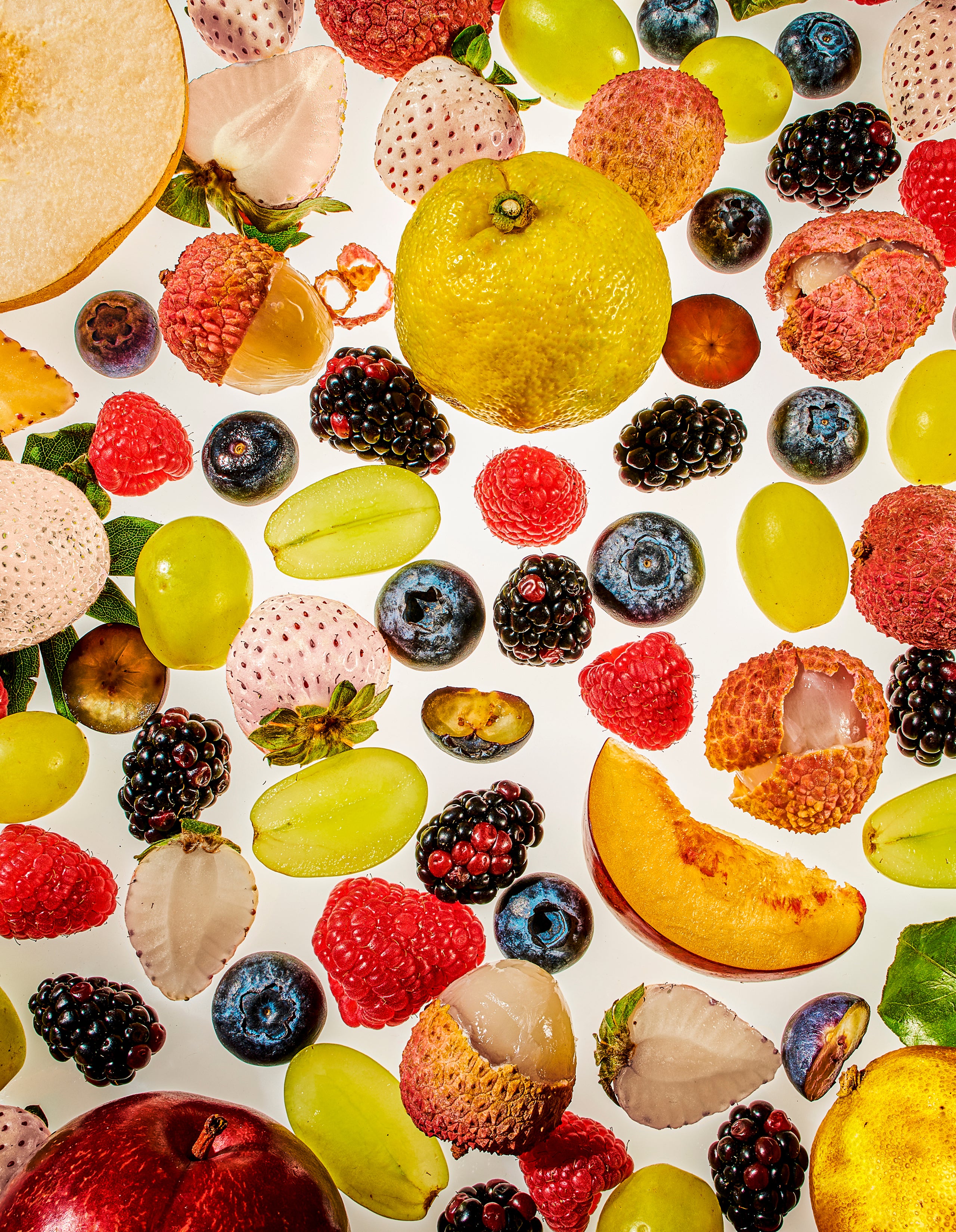 A vibrant, high-contrast flat-lay pattern of fresh fruits against a white background, featuring raspberries, blueberries, blackberries, halved green grapes, peeled lychees, a peach slice, and a whole yellow citrus fruit.