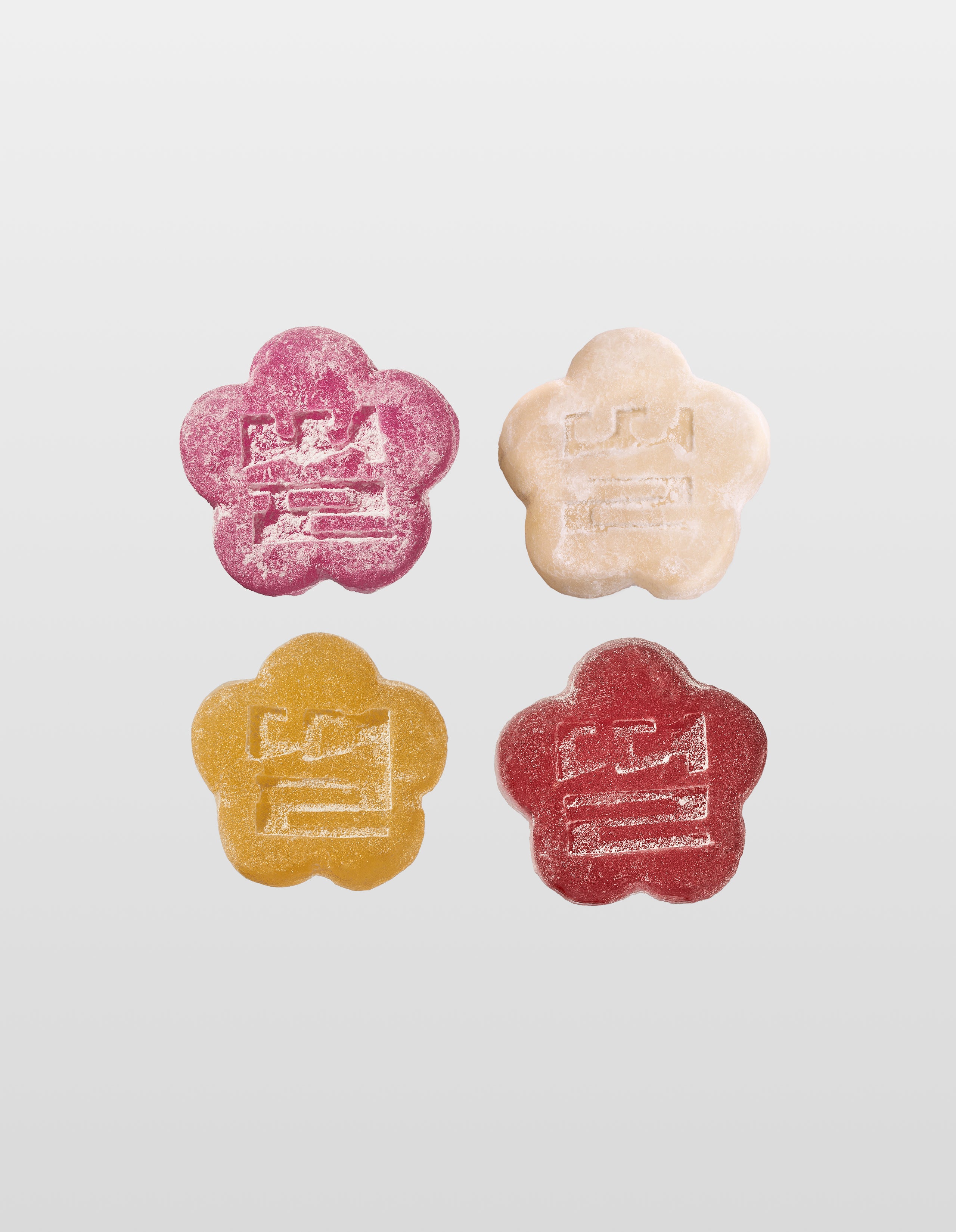 An overhead 2x2 grid displaying four flower-shaped mochi gummies, each labeled with dotted arrows: Lychee Dragon (deep pink), Yogurt (cream), Golden Pear (golden yellow), and Slumberberry (dark red). Each sugar-dusted gummy features a centered traditional emblem stamp.