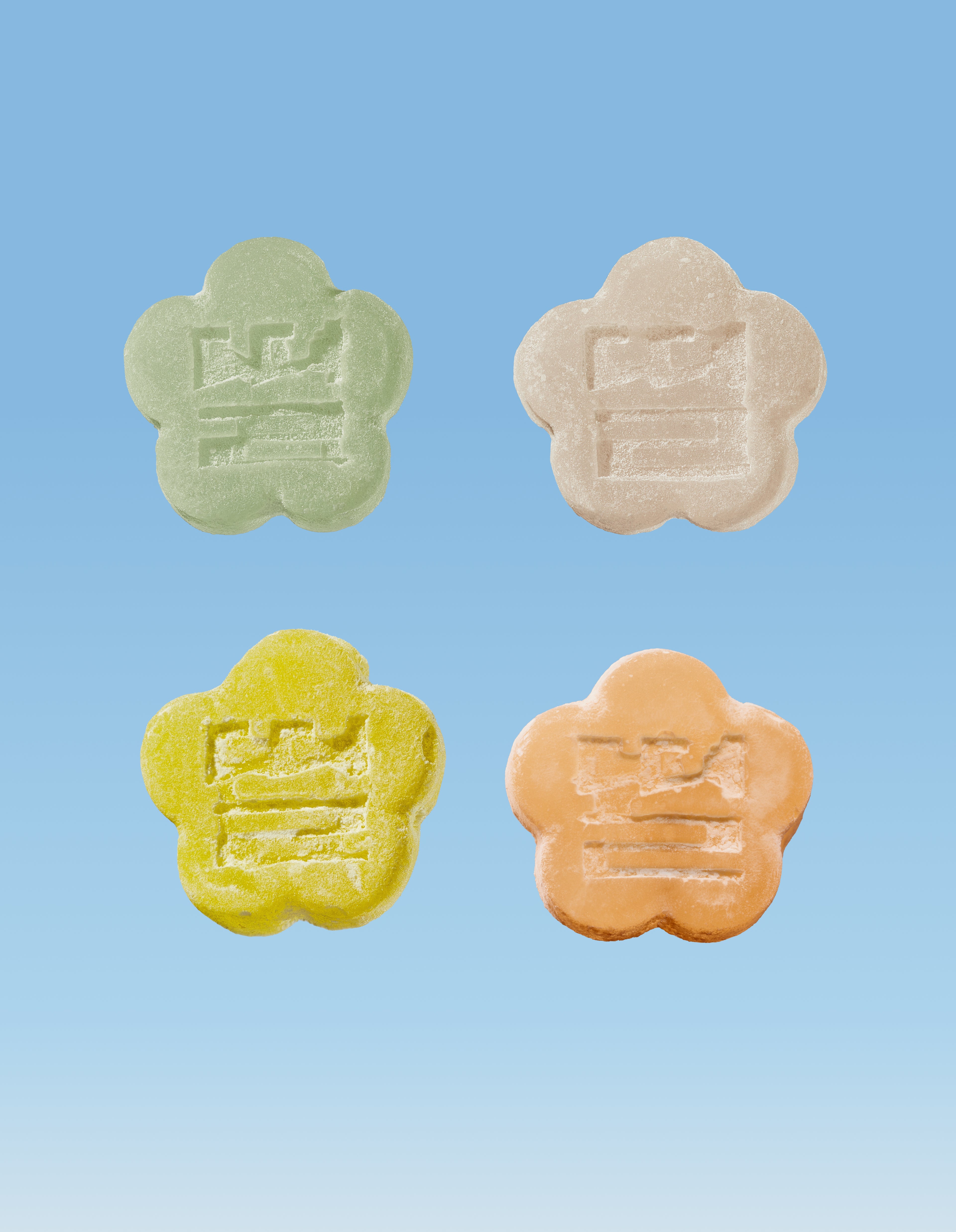Four colorful  flower-shaped Mochi Gummies in White, Yellow, Orange and Green on a blue background
