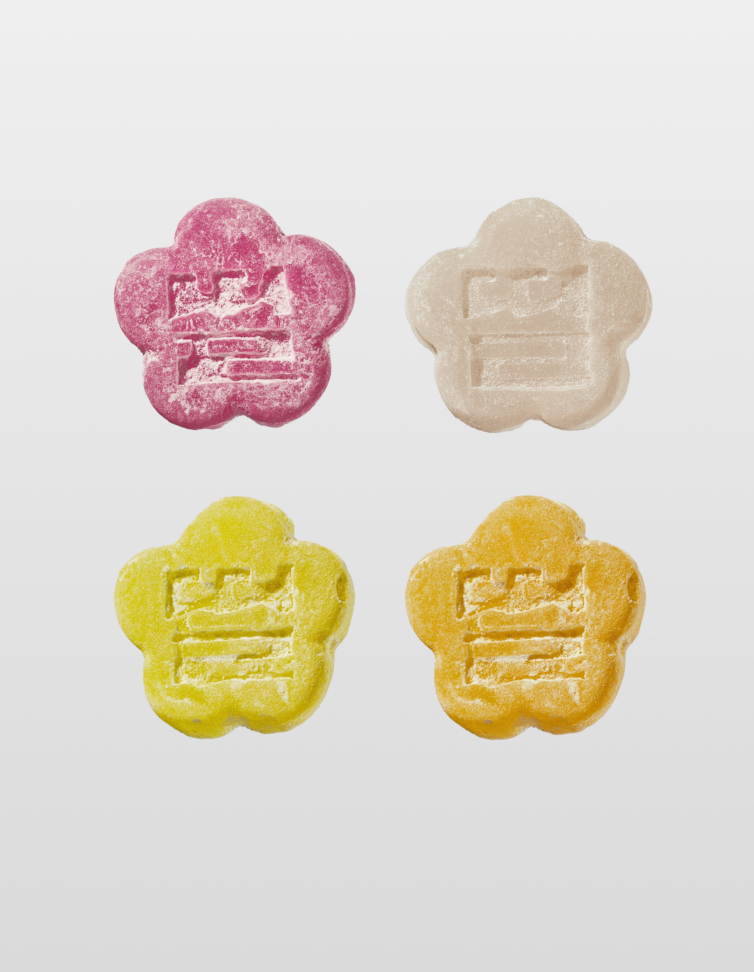 Four flower-shaped mochi gummies in pink, yellow, orange and white on a light gray background