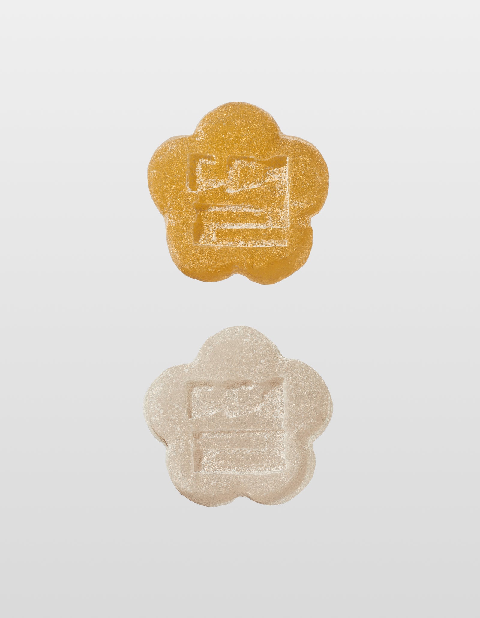 Two, yellow and white, flower-shaped mochi gummies with embossed 'ddul' korean character on a light gray background