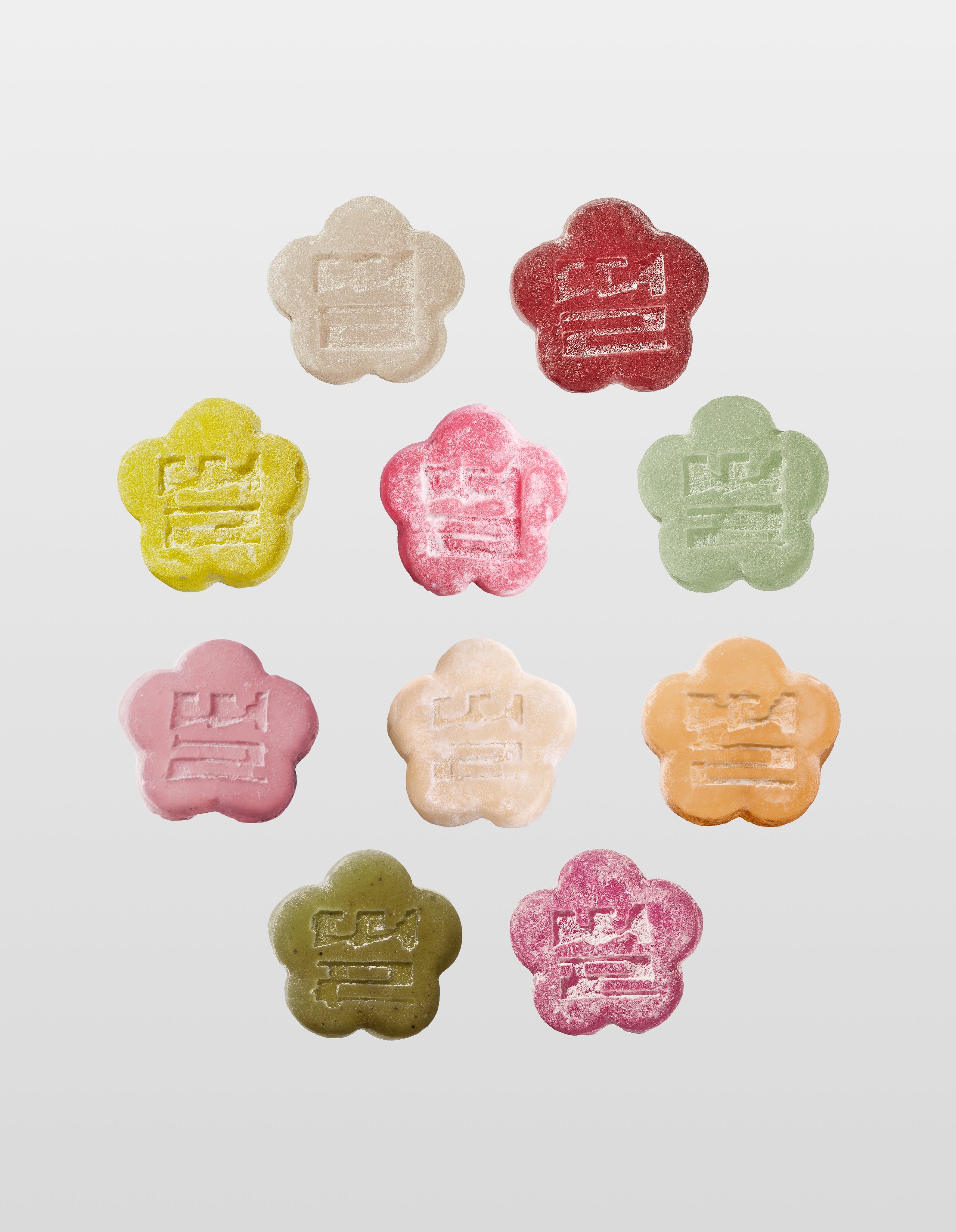 An overhead shot of ten flower-shaped mochi gummies arranged in a neat grid pattern against a light gray background. Each gummy is a different color—representing various fruit and tea flavors—and features a light sugar coating with an embossed traditional stamp in the center.
