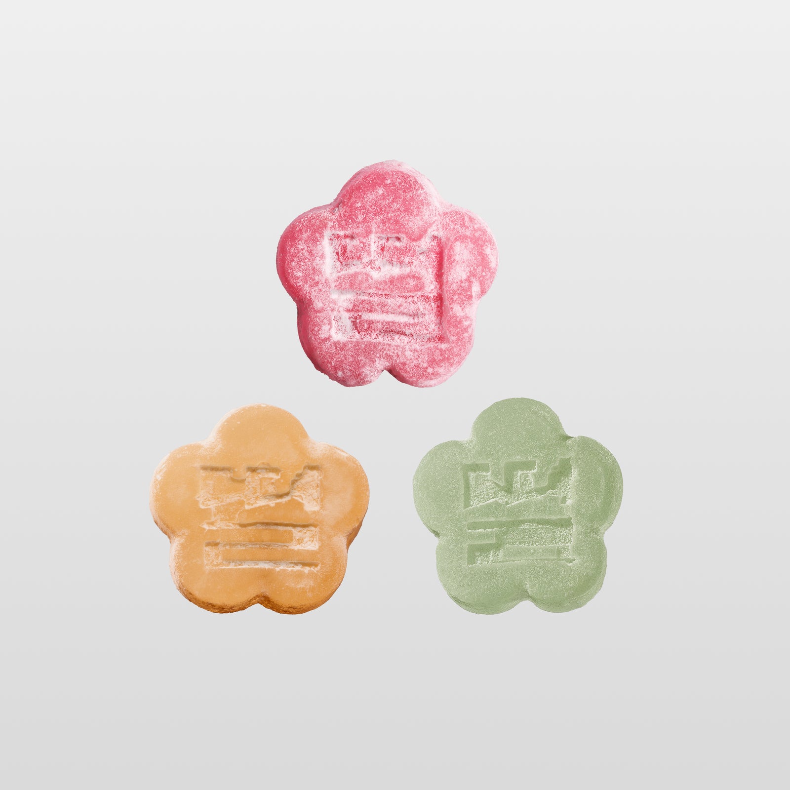 An overhead shot of three flower-shaped mochi gummies arranged in a neat grid pattern against a light gray background. Each gummy is a different color—representing various fruit flavors—and features a light sugar coating with an embossed traditional stamp in the center.