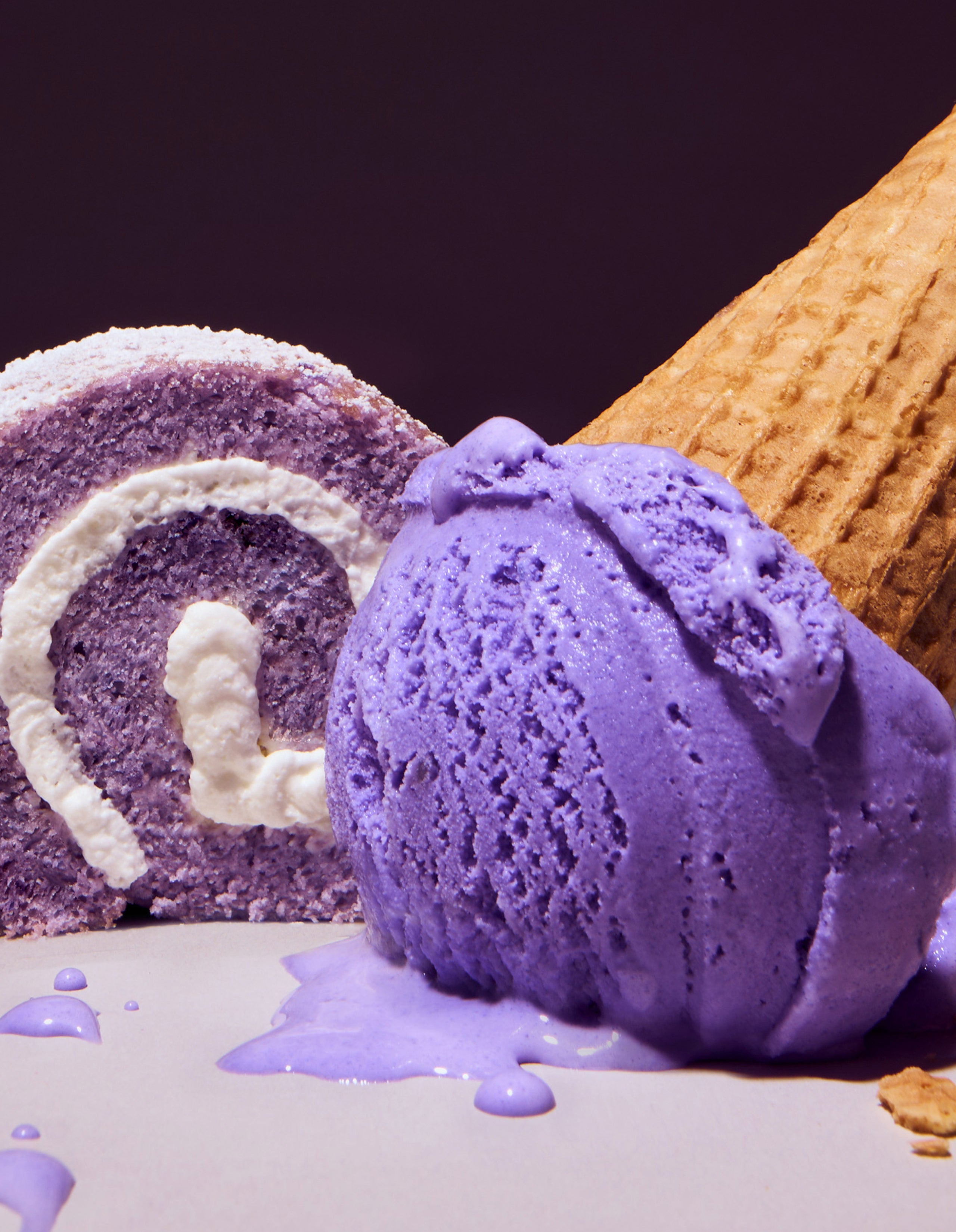 A lush close-up of a bright purple ube ice cream scoop melting slightly next to a slice of ube cake roll with white cream filling, with a waffle cone visible in the background to highlight the flavor profile.