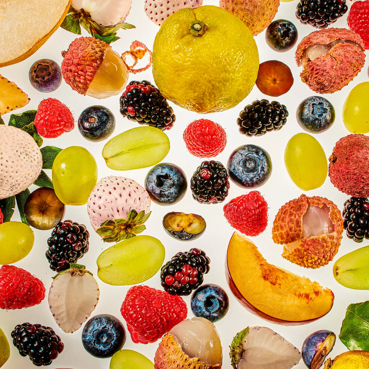 A vibrant, high-contrast flat-lay pattern of fresh fruits against a white background, featuring raspberries, blueberries, blackberries, halved green grapes, peeled lychees, a peach slice, and a whole yellow citrus fruit.