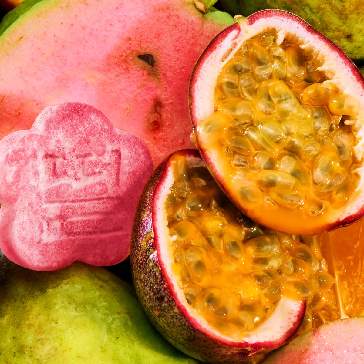 A vibrant, macro top-down shot of fresh ingredients featuring sliced pink guava and juicy halved passion fruits filled with golden seeds. A single flower-shaped pink mochi gummy is nestled among the fresh fruit on the left.