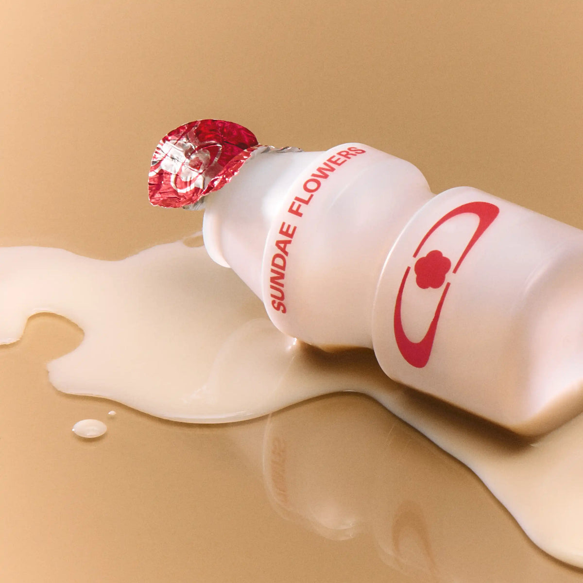 A macro shot of a small, white Asian-style probiotic yogurt bottle lying on its side on a warm tan surface, spilling a pool of creamy liquid. The bottle features a peeled red foil top and red text reading "SUNDAE FLOWERS."