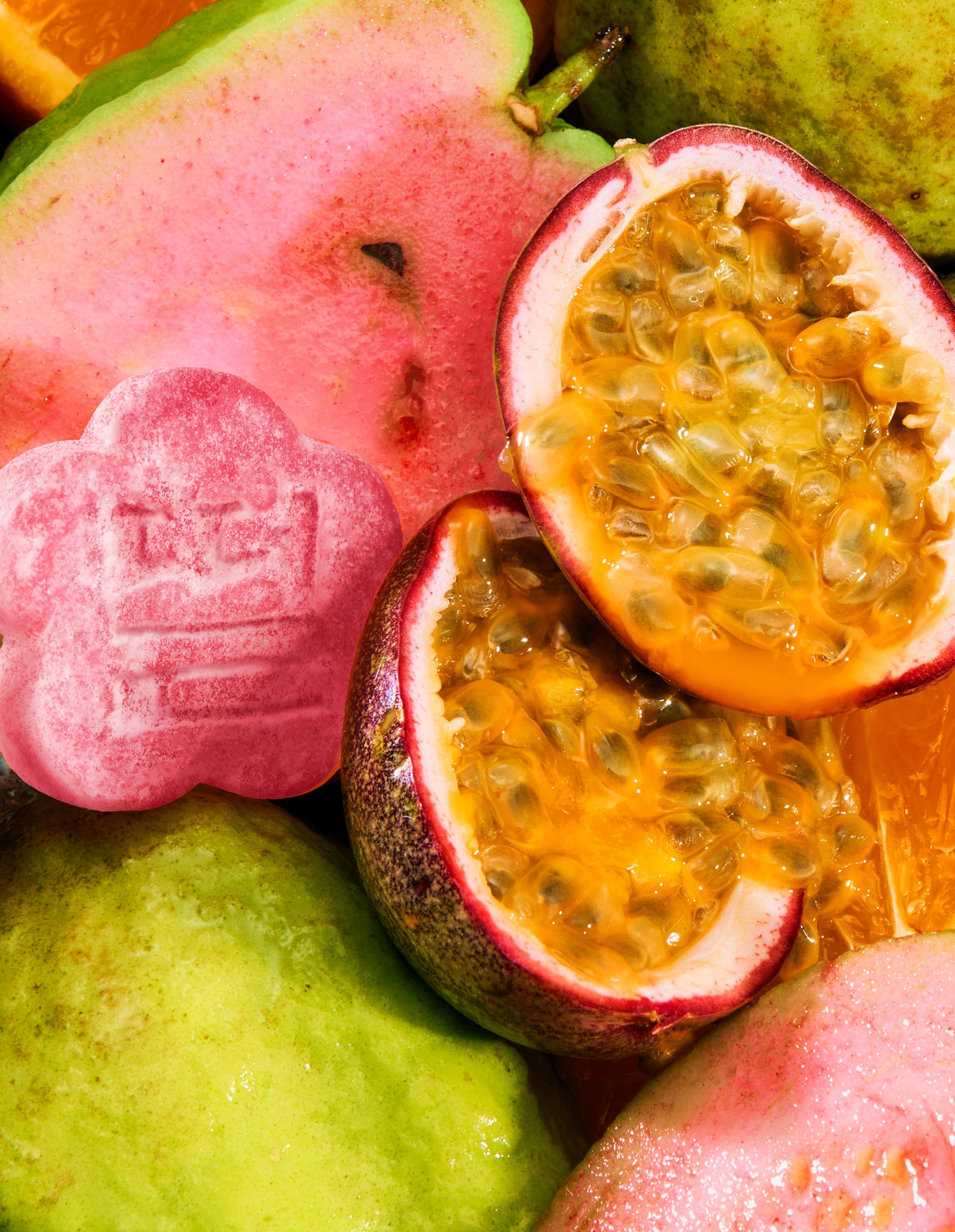 A vibrant, macro top-down shot of fresh ingredients featuring sliced pink guava and juicy halved passion fruits filled with golden seeds. A single flower-shaped pink mochi gummy is nestled among the fresh fruit on the left.