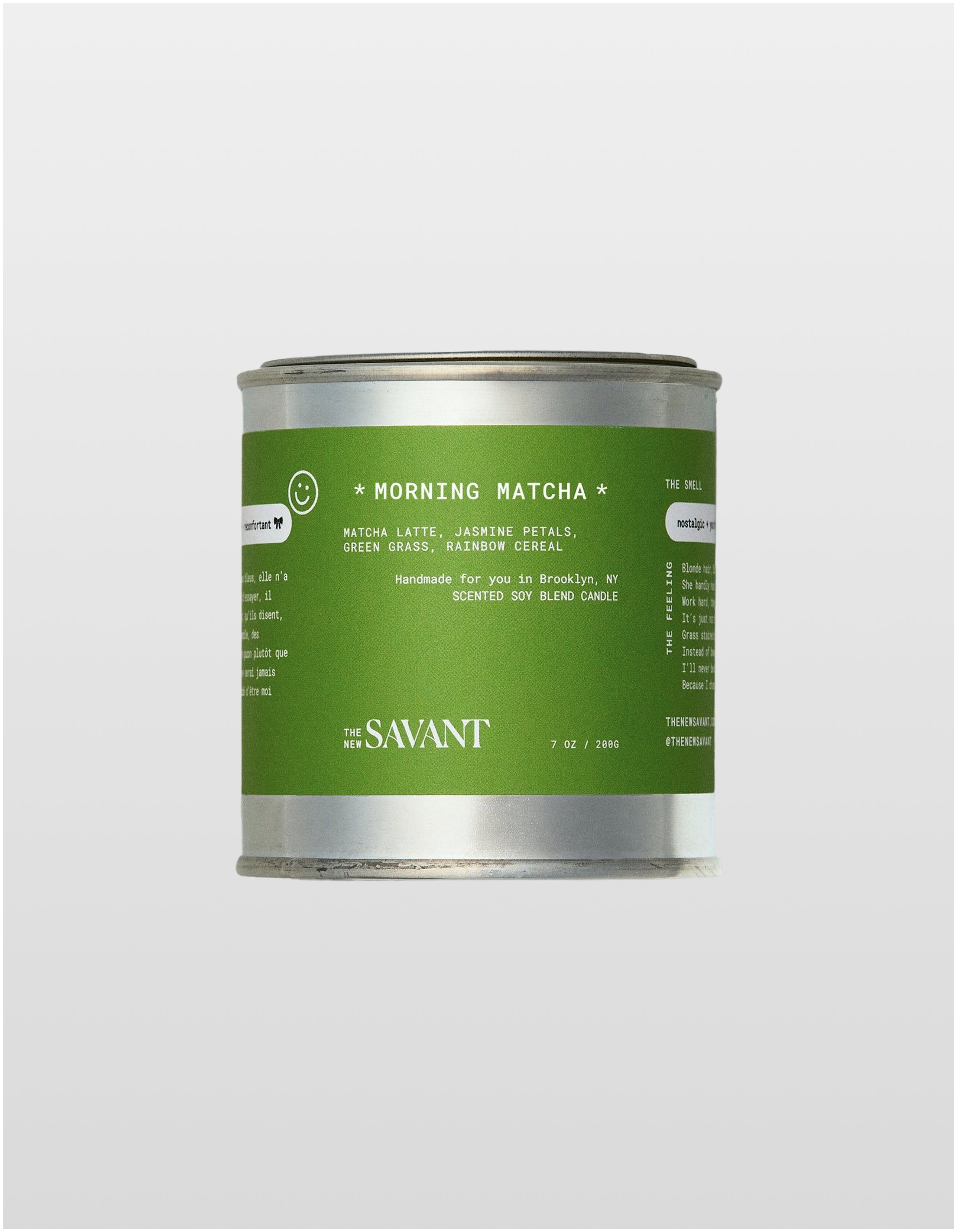 A product shot of a silver metal tin candle by The New Savant, centered against a minimal gray background. The candle has a vibrant green label titled 