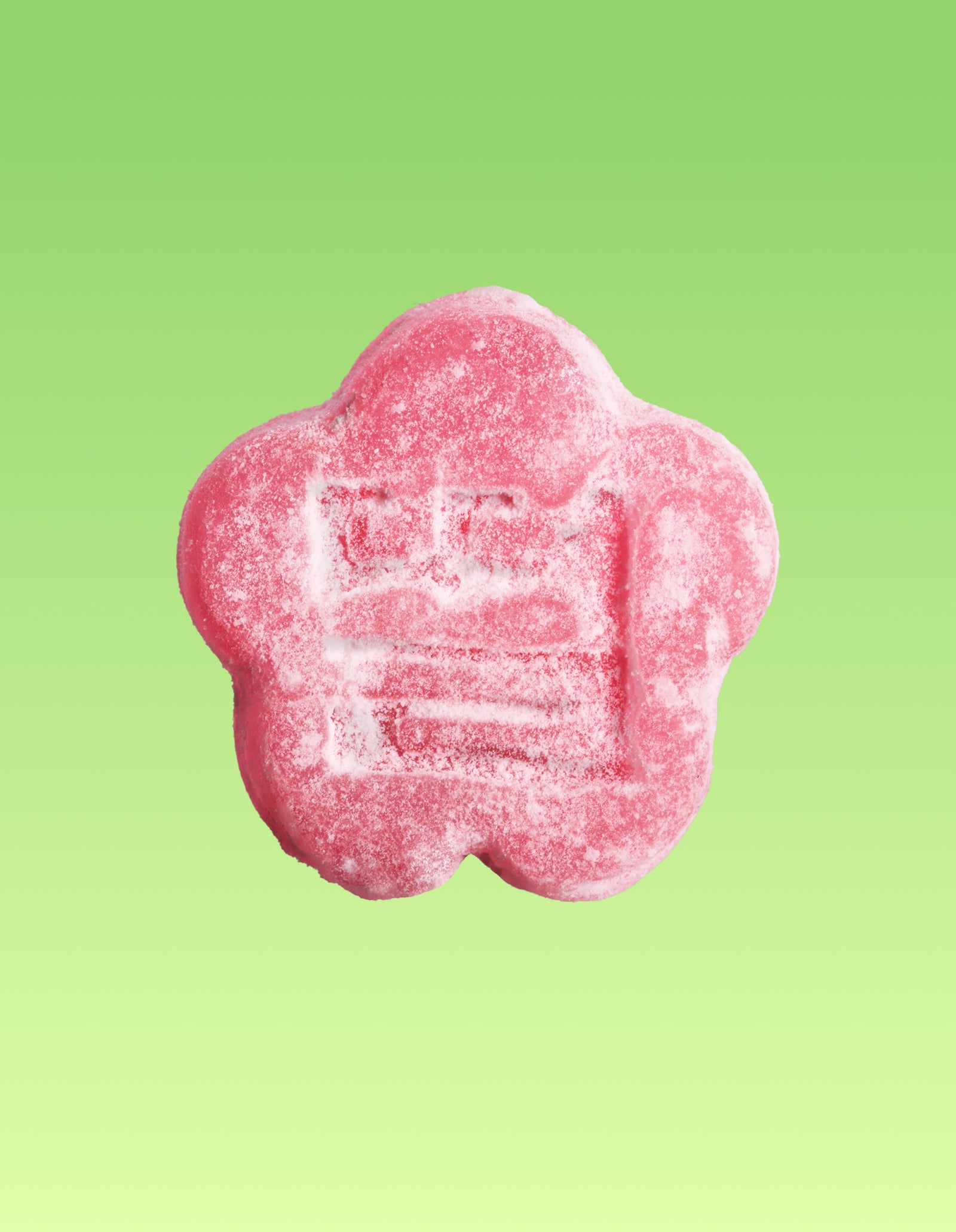 A close-up of a single pink, flower-shaped mochi gummy lightly coated in sugar crystals. The center of the gummy features an embossed traditional stamp, all set against a clean lime green gradient background.