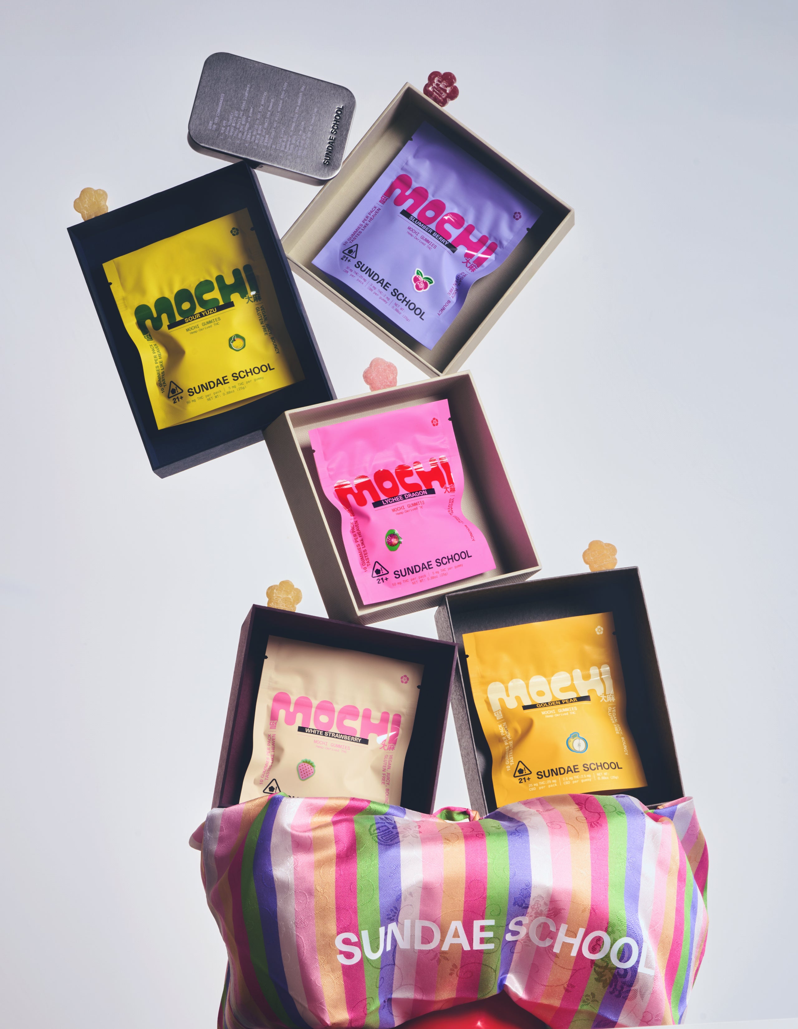 An Image of 10ct Slumber Berry Mochi gummy, Sour Yuzu Mochi gummy, Lychee Dragon Mochi gummy, Golden Pear Mochi gummy, White Strawberry Mochi Gummy packagings in a resealable bag form individually put in a box along with Mochi gummies, Sundae School Mochi Tin, and Sundae School mini Bojagi Bag. 