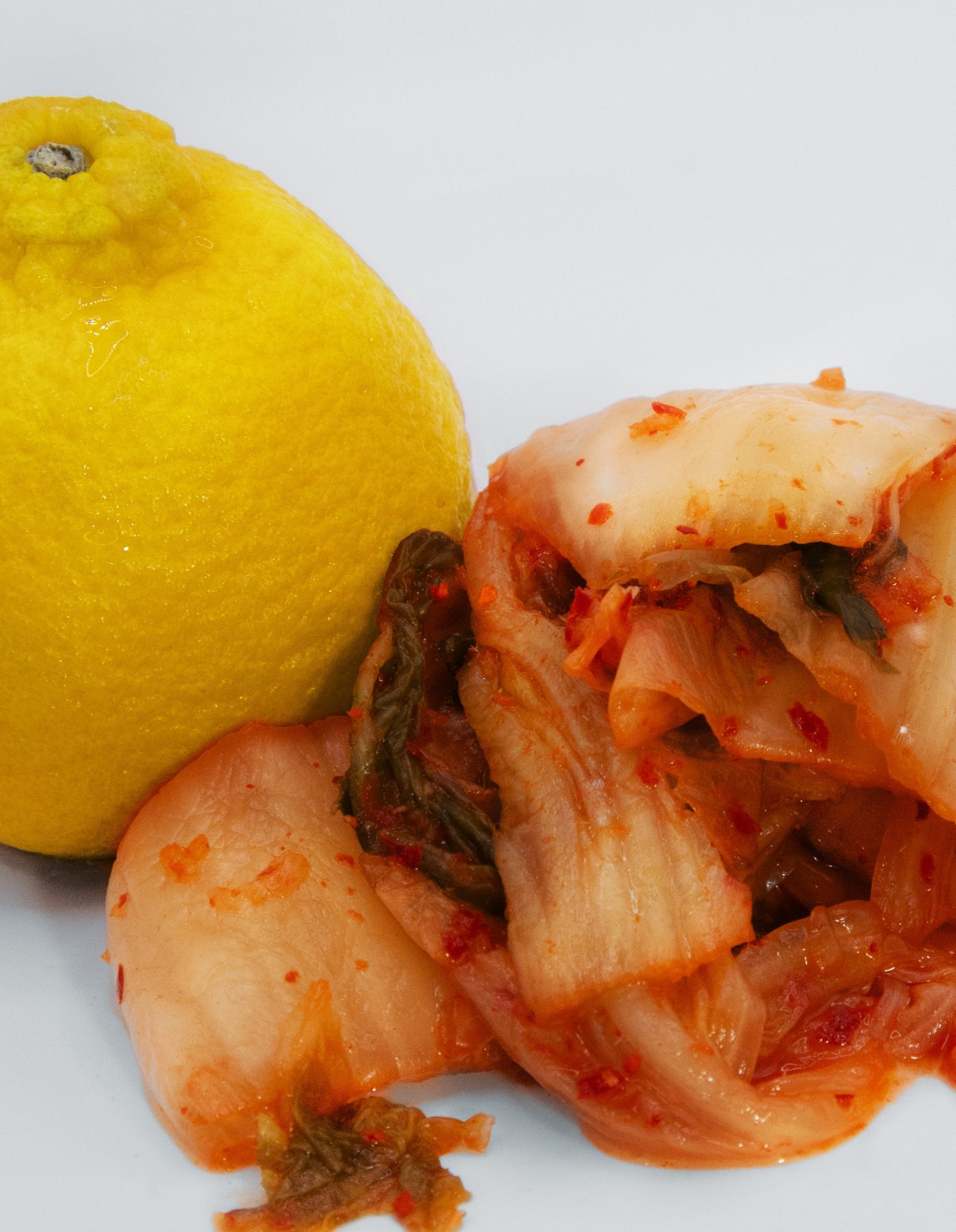 A close-up image of a yuzu on the left and pile of kimchee to the right on a white surface. 
