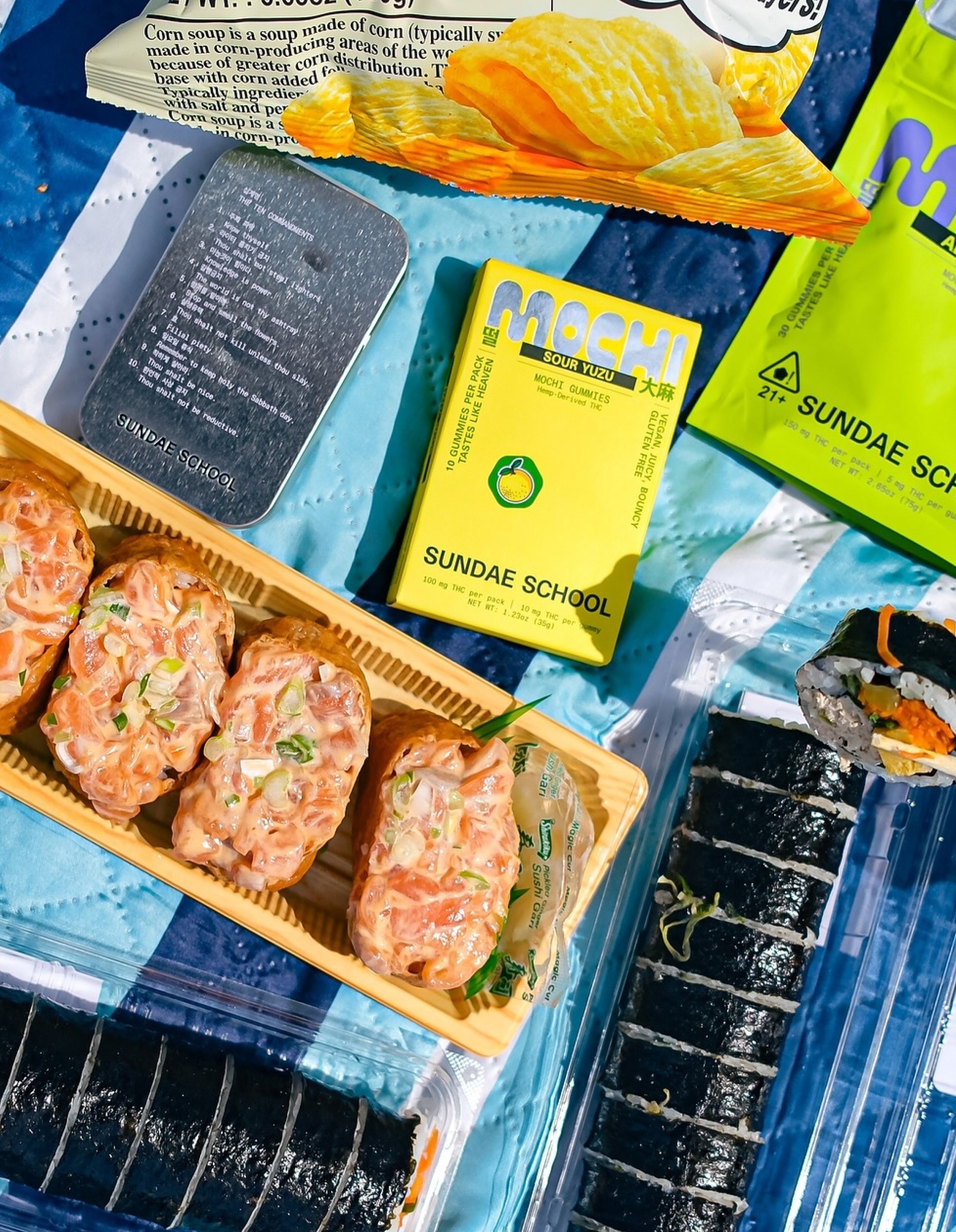 Assorted snacks including two types of sushi, a package of korean turtle chips, a tin of sour yuzu sundae school edibles, and a mylar bag of sundae school aloe grape edibles on a blue checkered picnic blanket.