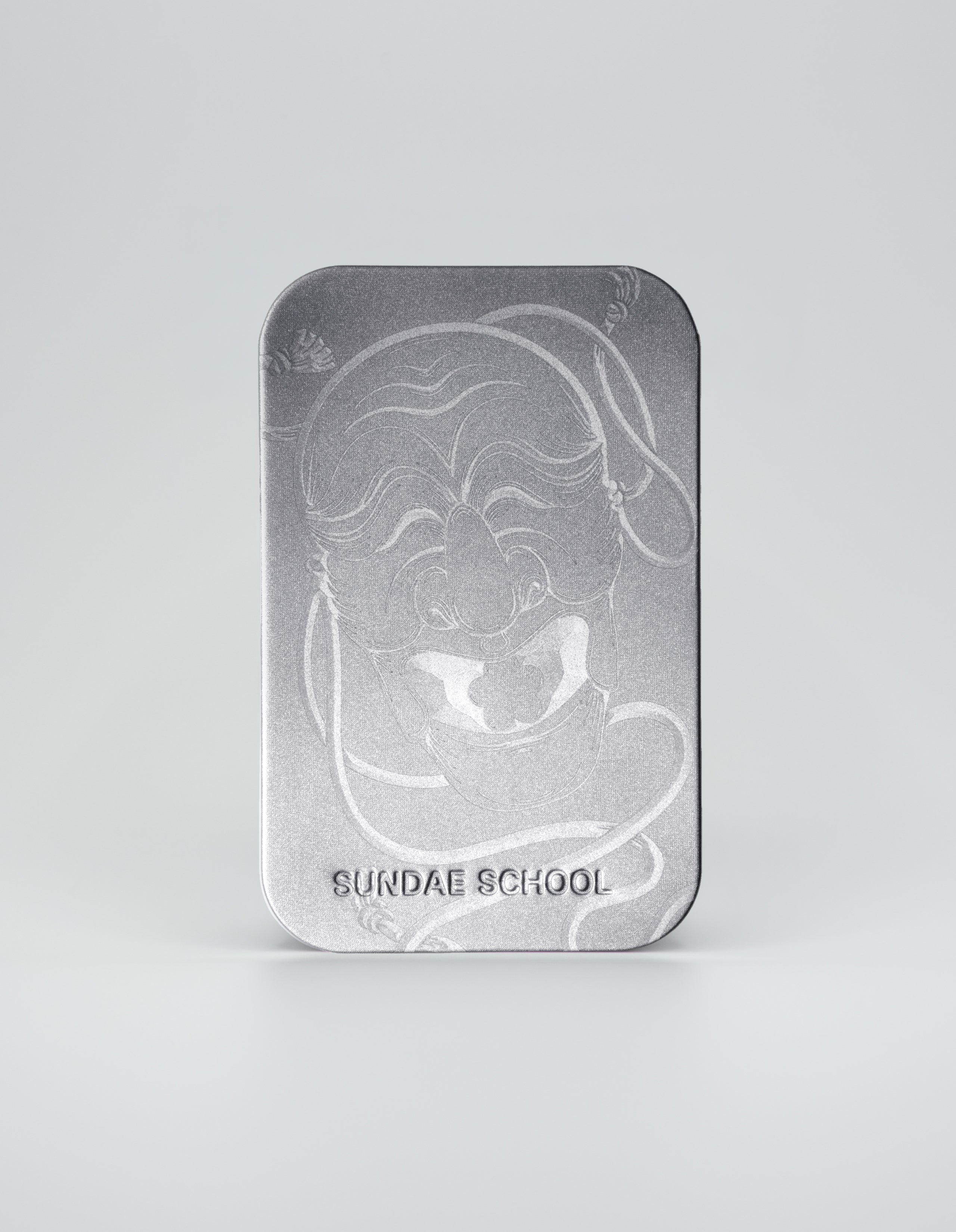 A close-up of a vertical silver metallic tin with a detailed etching of a traditional smiling Korean Hahoe mask. The words 